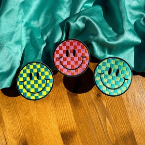 May include: Three embroidered smiley face patches with a checkered pattern. The patches are yellow and green, red and pink, and blue and green.