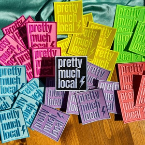 May include: A collection of colorful embroidered patches with the text "pretty much local" and a lightning bolt design. The patches are arranged in a random pattern on a wooden surface.