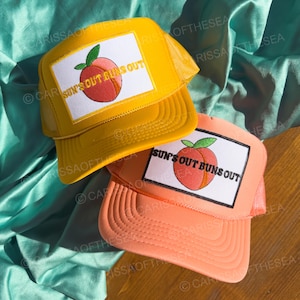 Op de afbeelding: Twee trucker caps met een perzikgrafiek en de tekst "Sun's Out Buns Out". De ene pet is geel en de andere is perzikkleurig.