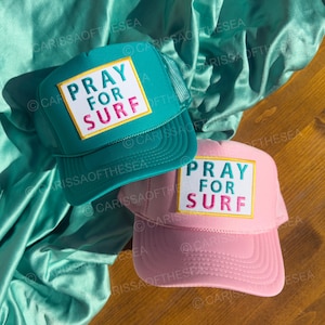 Könnte beinhalten: Zwei Trucker-Caps mit Netzrücken. Die Caps sind grün und pink. Beide Caps haben einen weißen Patch mit dem Text "Pray for Surf" in pinken und gelben Buchstaben.