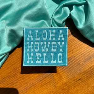 May include: A teal blue fabric patch with white embroidered text that reads "Aloha Howdy Hello".