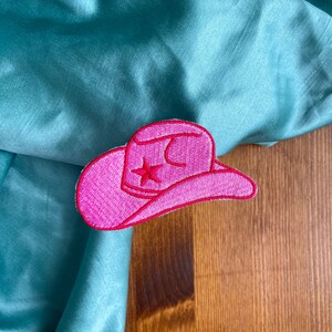 May include: Pink felt cowboy hat patch with a white star on a teal fabric background.