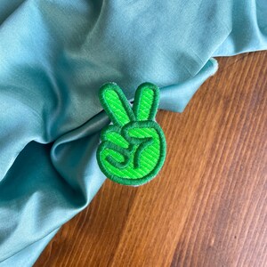 K&ouml;nnte beinhalten: Gr&uuml;ner gestickter Peace-Zeichen-Patch. Der Patch befindet sich auf einem hellblauen Stoffhintergrund.