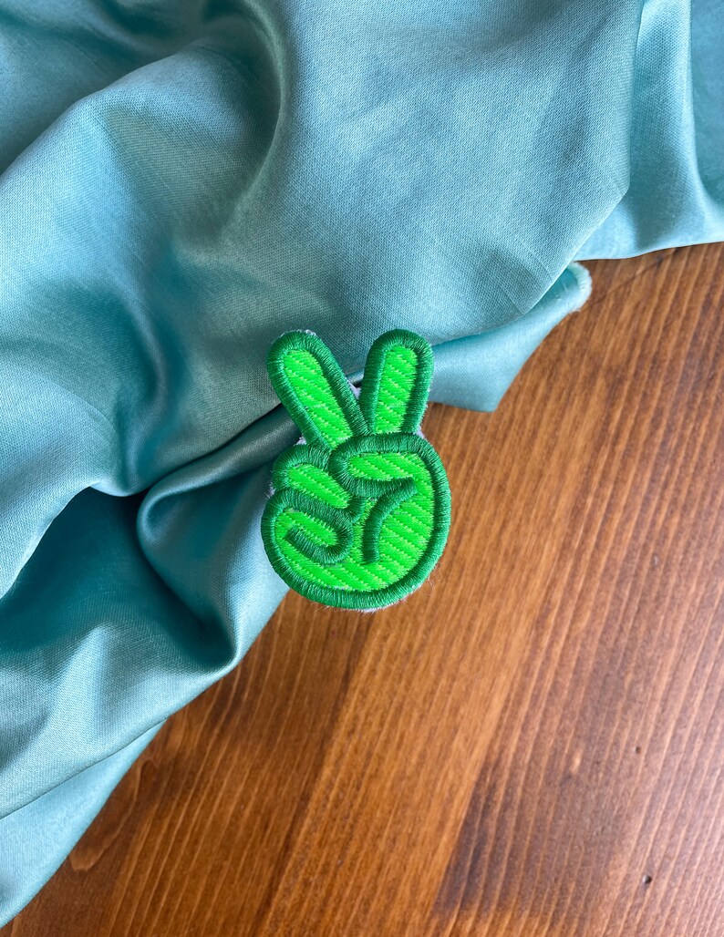 K&ouml;nnte beinhalten: Gr&uuml;ner gestickter Peace-Zeichen-Patch. Der Patch befindet sich auf einem hellblauen Stoffhintergrund.