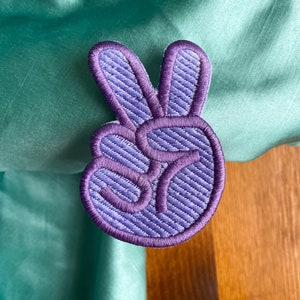 K&ouml;nnte beinhalten: Lilafarbener gestickter Peace-Zeichen-Patch mit strukturiertem Stoff.