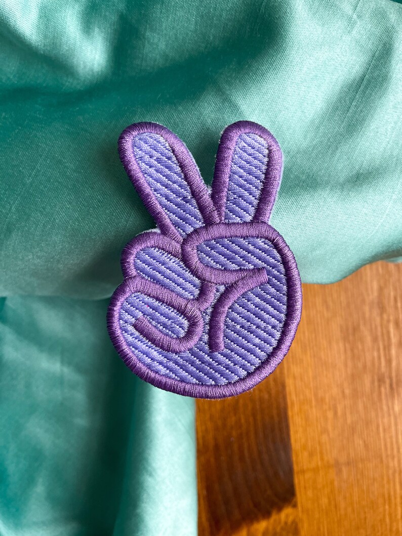 K&ouml;nnte beinhalten: Lilafarbener gestickter Peace-Zeichen-Patch mit strukturiertem Stoff.