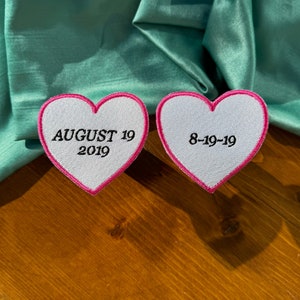 May include: Two white heart-shaped patches with pink outlines. The left patch has the text "AUGUST 19 2019" embroidered on it. The right patch has the text "8-19-19" embroidered on it.