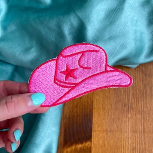 May include: Pink felt cowboy hat patch with a white star in the center. The patch is embroidered with white thread.