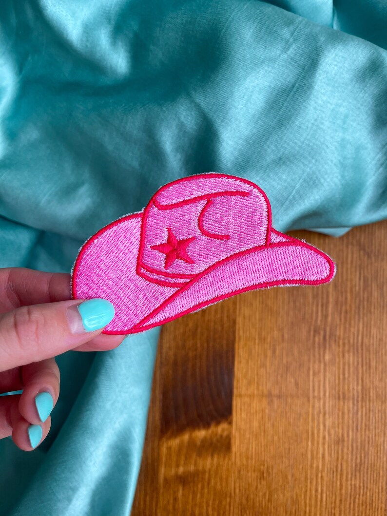 May include: Pink felt cowboy hat patch with a white star in the center. The patch is embroidered with white thread.