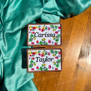 May include: Two embroidered patches with floral borders and white backgrounds. The top patch says "Carissa" and the bottom patch says "Taylor".