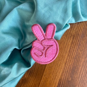K&ouml;nnte beinhalten: Rosa gestickter Peace-Zeichen-Patch. Der Patch befindet sich auf einem t&uuml;rkisfarbenen Stoffhintergrund.