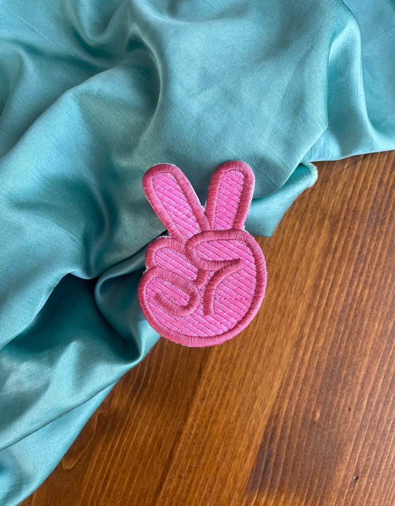 K&ouml;nnte beinhalten: Rosa gestickter Peace-Zeichen-Patch. Der Patch befindet sich auf einem t&uuml;rkisfarbenen Stoffhintergrund.