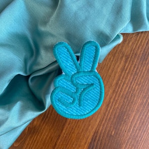 K&ouml;nnte beinhalten: Ein gestickter Peace-Zeichen-Patch in blaugr&uuml;n. Der Patch ist aus weichem, strukturiertem Stoff gefertigt und hat ein erhabenes Design.