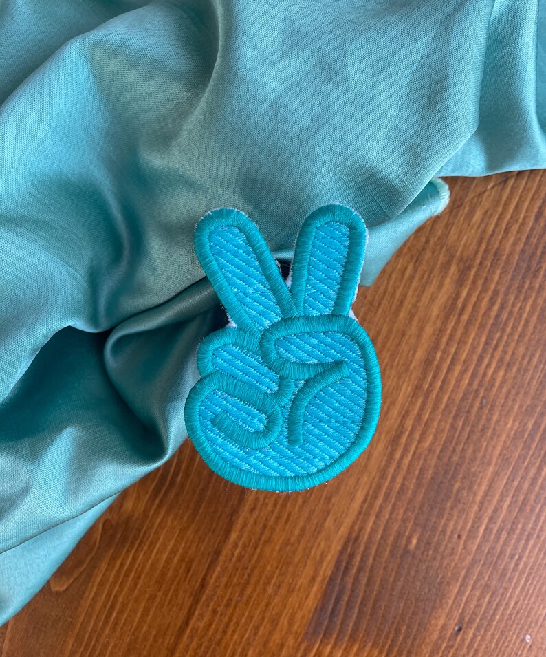 K&ouml;nnte beinhalten: Ein gestickter Peace-Zeichen-Patch in blaugr&uuml;n. Der Patch ist aus weichem, strukturiertem Stoff gefertigt und hat ein erhabenes Design.