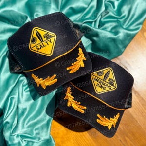 Puede incluir: Dos gorras de camionero negras con ribetes dorados y diseños bordados. Una gorra presenta un diamante amarillo con las palabras "Salty Sea" y un gráfico de tabla de surf. La otra gorra presenta un diamante amarillo con las palabras "Mermaid Xing" y un gráfico de tabla de surf.