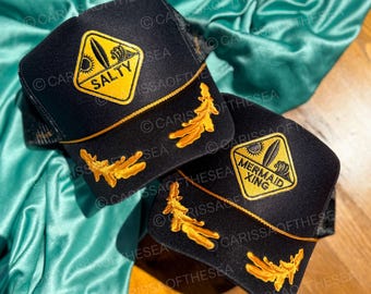 May include: Two black trucker hats with gold trim and embroidered designs. One hat features a yellow diamond with the words "Salty Sea" and a surfboard graphic. The other hat features a yellow diamond with the words "Mermaid Xing" and a surfboard graphic.