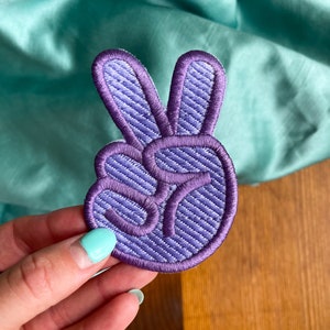 K&ouml;nnte beinhalten: Lilafarbener gestickter Peace-Zeichen-Patch mit strukturiertem Stoff.