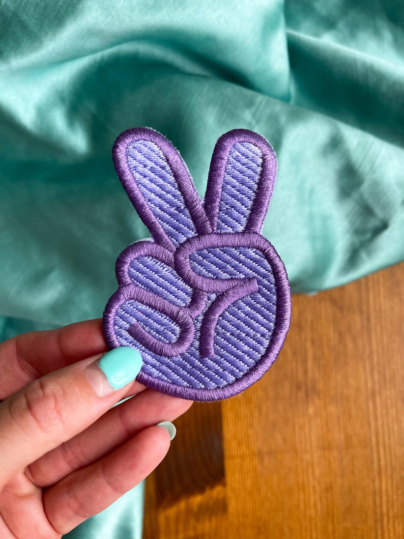 K&ouml;nnte beinhalten: Lilafarbener gestickter Peace-Zeichen-Patch mit strukturiertem Stoff.