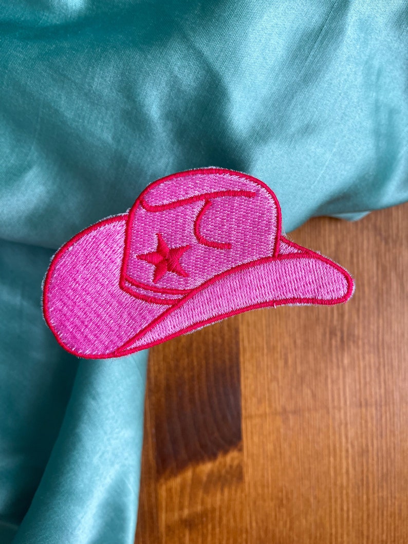 May include: Pink felt cowboy hat with a white star embroidered on the front. The hat is on a wooden surface.