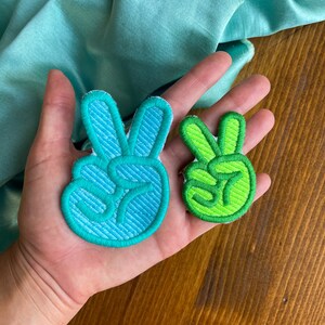 K&ouml;nnte beinhalten: Zwei Peace-Zeichen-Patches, einer in blaugr&uuml;n und einer in gr&uuml;n, werden auf einer Hand gezeigt. Die Patches sind mit einem Peace-Zeichen bestickt.