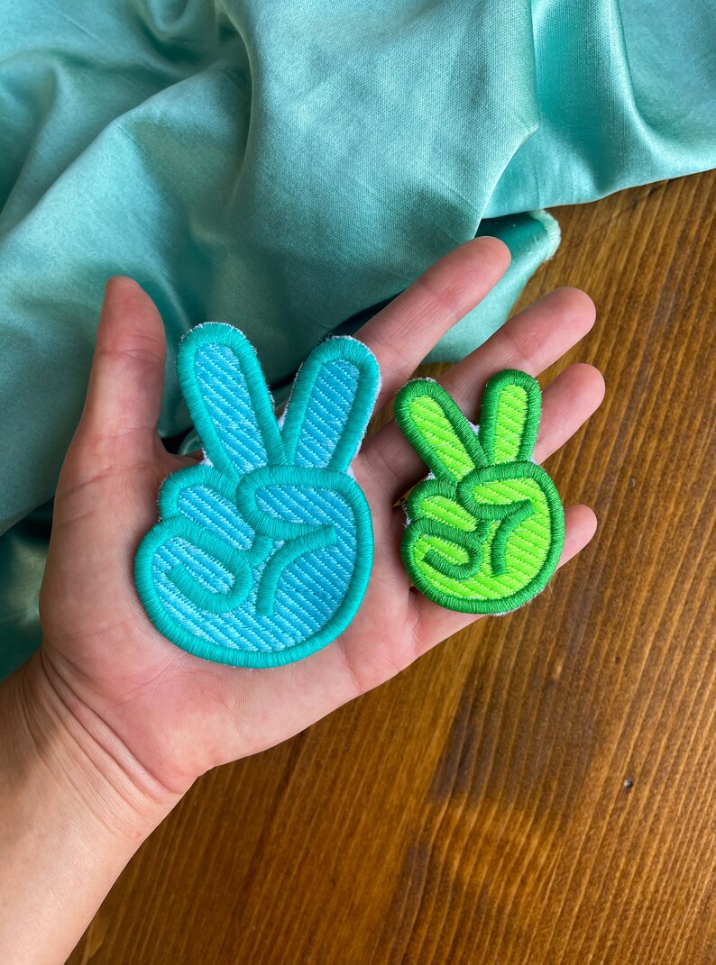 K&ouml;nnte beinhalten: Zwei Peace-Zeichen-Patches, einer in blaugr&uuml;n und einer in gr&uuml;n, werden auf einer Hand gezeigt. Die Patches sind mit einem Peace-Zeichen bestickt.