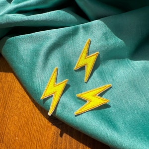 May include: Three yellow lightning bolt iron-on patches on a teal fabric background.