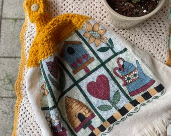 Op de afbeelding: Een witte keukendoek met een gele gehaakte lusvormige handgreep. De handdoek heeft een kleurrijk patchwork-ontwerp met afbeeldingen van een vogelhuisje, een bijenkorf, een hart en een gieter. De handdoek heeft een franje rand.