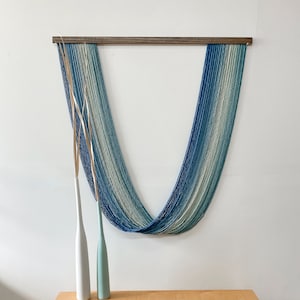May include: A wall hanging made of blue and white yarn, with a wooden dowel at the top. The hanging is in the shape of a U, and is hanging on a white wall. There are two white vases on a wooden bench in front of the hanging.