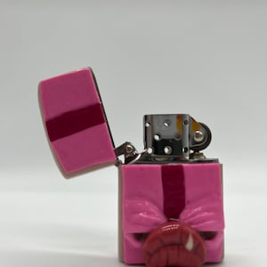 Può includere: Un accendino rosa e rosso a forma di testa di maiale. L'accendino è aperto e il coperchio è ribaltato all'indietro. L'accendino ha una cerniera argentata e una ruota argentata sul lato.