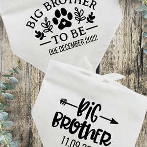 May include: Two white fabric bibs with black text. The top bib says "Big Brother - To Be - Due December 2022" with a paw print and floral designs. The bottom bib says "Big Brother - 11.09.22" with an arrow.