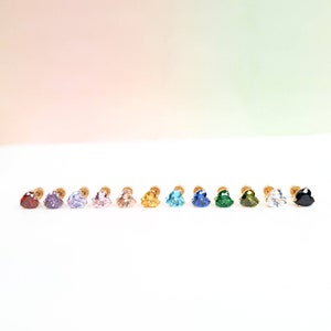 Puede incluir: Un conjunto de 12 pendientes de botón en forma de corazón con piedras preciosas de diferentes colores. Los pendientes están dispuestos en una fila sobre una superficie blanca. Los colores de las piedras preciosas son rojo, morado, morado claro, rosa, amarillo, azul claro, azul, verde, verde oscuro, transparente y negro.