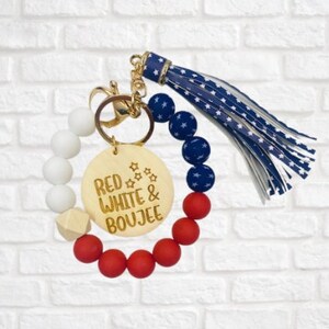 May include: Red, white, and blue beaded keychain with a wooden charm that says "Red White & Boujee". The keychain has a gold key ring and a blue and white star patterned tassel.