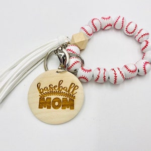 Puede incluir: Llavero con un encanto de béisbol blanco y una etiqueta de madera que dice "baseball mom". El llavero tiene un borla blanca.
