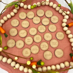 May include: A set of 20 wooden tokens with various rewards written on them, such as "1 day trip of choice", "$5", "Cook an item of choice", "Ice cream sundae night", and "You pick dinner". The tokens are arranged on a pink surface with a white bead garland around the edge.