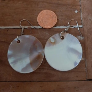 May include: A pair of round, iridescent earrings with silver hooks. The earrings are a light cream colour with brown and white marbling. A copper coin is next to the earrings.