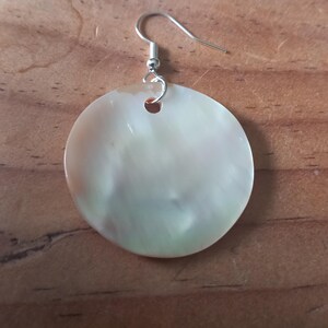 May include: A circular earring made of iridescent mother-of-pearl with a smooth, polished surface. The disc features a small hole at the top, connected to a silver-toned metal fishhook ear wire for hanging. The material displays subtle white, cream, and pale green colour variations.