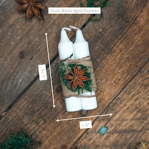 May include: Two white spell candles, 3.75 inches tall and 1.3 inches wide, are wrapped with twine, moss, and a star anise. The label reads "Plain White Spell Candles". The candles are on a rustic wooden surface.