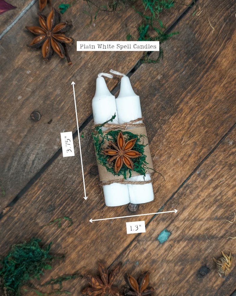 May include: Two white spell candles, 3.75 inches tall and 1.3 inches wide, are wrapped with twine, moss, and a star anise. The label reads "Plain White Spell Candles". The candles are on a rustic wooden surface.