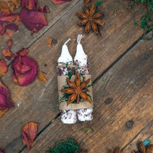 May include: Two white candles wrapped in a brown paper with dried flowers and star anise. The candles are on a rustic wooden surface, surrounded by dried rose petals, moss, and more star anise.