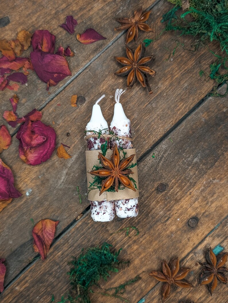 May include: Two white candles wrapped in a brown paper with dried flowers and star anise. The candles are on a rustic wooden surface, surrounded by dried rose petals, moss, and more star anise.