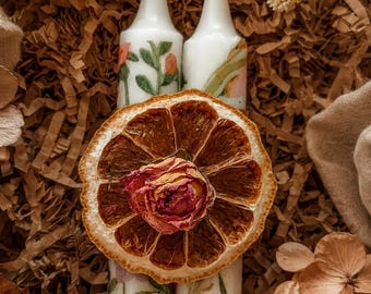 Peut inclure: Deux bougies effilées blanches décorées de motifs floraux avec des tiges vertes et des fleurs orange. Une grande tranche d'orange séchée et circulaire est placée horizontalement au centre des bougies, surmontée d'un bouton de rose rose séché.