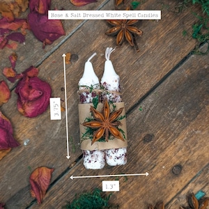 May include: Two white spell candles, 3.75 inches tall and 1.3 inches wide, are wrapped together with a brown paper band, dried roses, and star anise. The candles are decorated with rose petals and salt. The text on the image reads "Rose & Salt Dressed White Spell Candles."