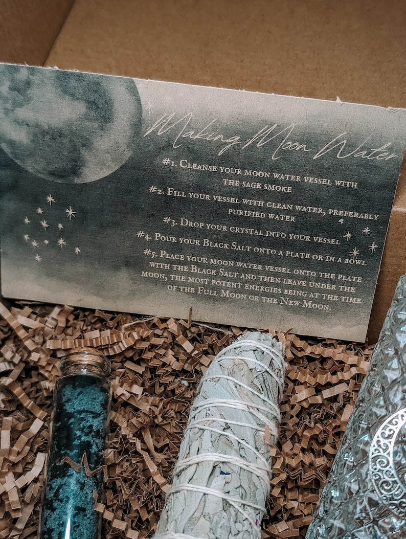 May include: A box with instructions for making moon water. The box contains a white sage smudge stick, a small glass bottle with blue crystals, and a piece of paper with instructions on how to make moon water.