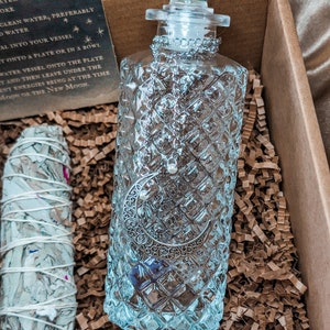 May include: A clear glass bottle with a silver crescent moon design and a glass stopper. The bottle is sitting in a box filled with brown packing paper. The bottle is labeled "Moon Water".