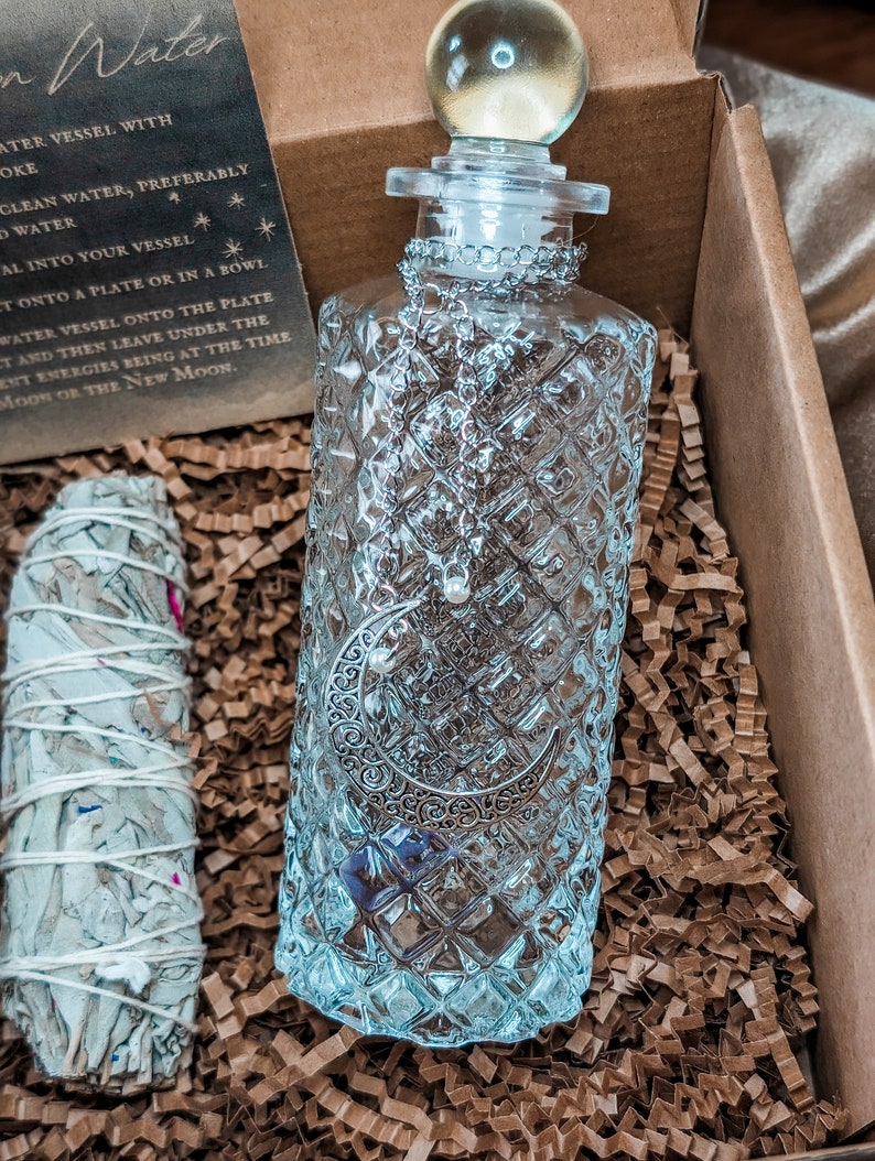 May include: A clear glass bottle with a silver crescent moon design and a glass stopper. The bottle is sitting in a box filled with brown packing paper. The bottle is labeled "Moon Water".
