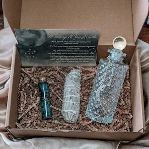 May include: A brown cardboard box with a white card that says "Making Moon Water" and instructions for making moon water. The box contains a clear glass bottle with a stopper, a small bottle of blue liquid, and a bundle of white sage.