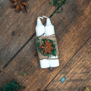 May include: Two white candles are bundled together with twine, brown paper, and green moss, and decorated with a star anise. The candles are on a rustic wooden surface, with additional star anise and moss scattered around.