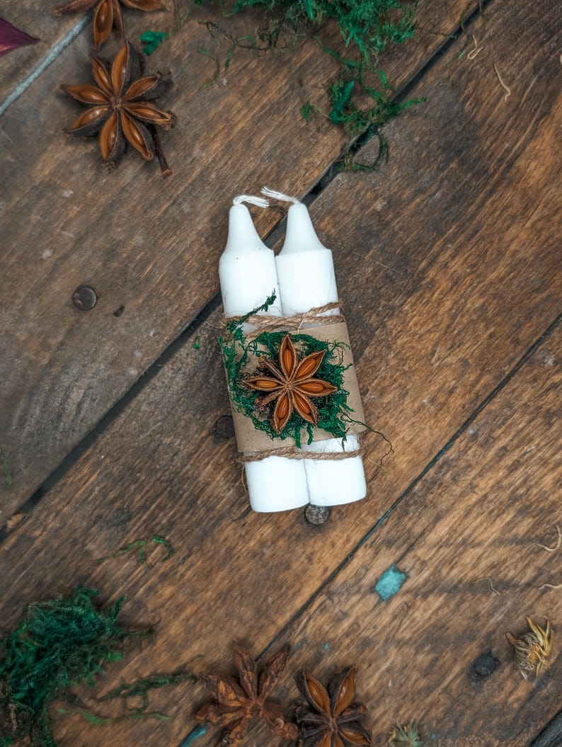 May include: Two white candles are bundled together with twine, brown paper, and green moss, and decorated with a star anise. The candles are on a rustic wooden surface, with additional star anise and moss scattered around.
