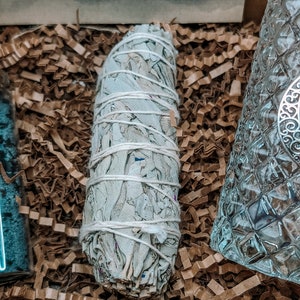 May include: A white sage smudge stick wrapped in white string, sitting on brown shredded paper. The sage is a light gray color and has a natural, earthy texture.