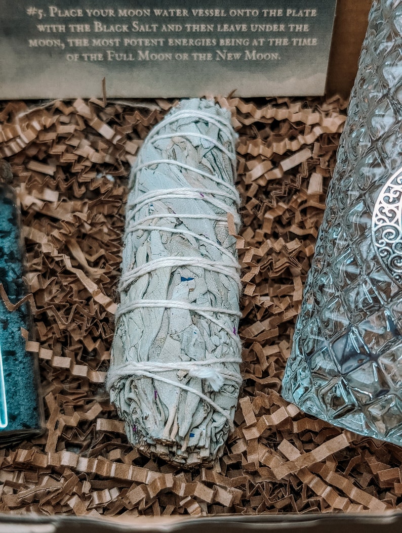 May include: A white sage smudge stick wrapped in white string, sitting on brown shredded paper. The sage is a light gray color and has a natural, earthy texture.